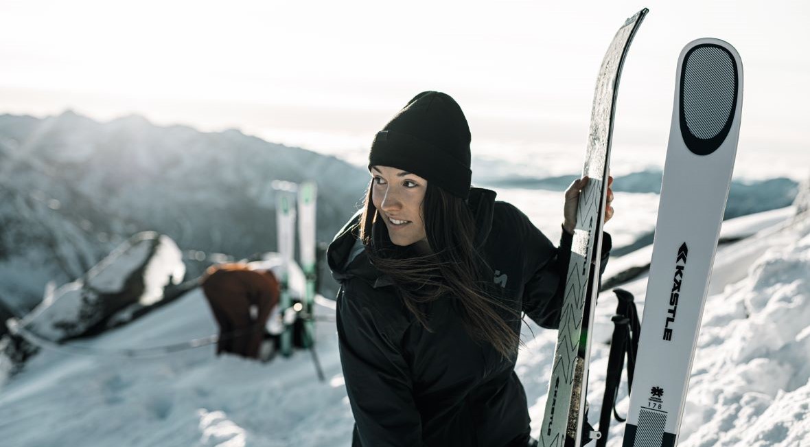 Eine Frau in einer schwarzen Jacke und Mütze steht lächelnd auf einem verschneiten Berggipfel mit einem Paar Ski, während die Sonne im Hintergrund die Berglandschaft beleuchtet.